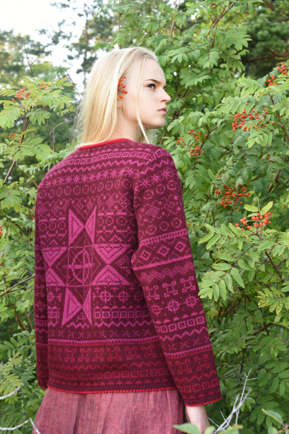 blonde model in front of a tree wearing a short red wool cardigan with power symbols from kauni design