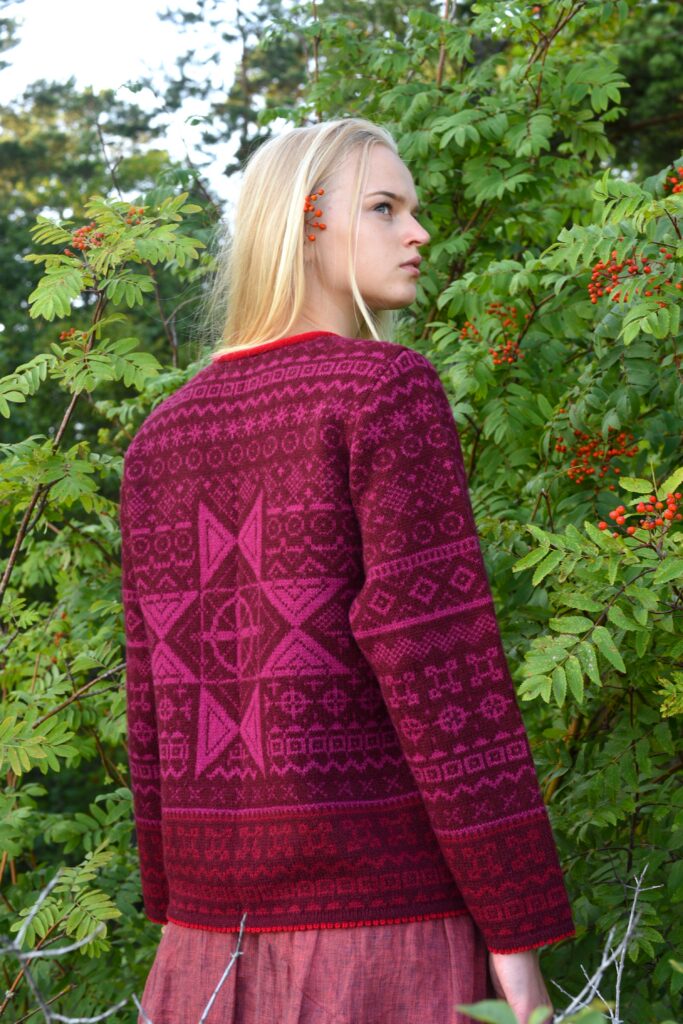 blonde woman standing in front of a rowan tree wearing a red patterned short wool cardigan with ancient power symbols