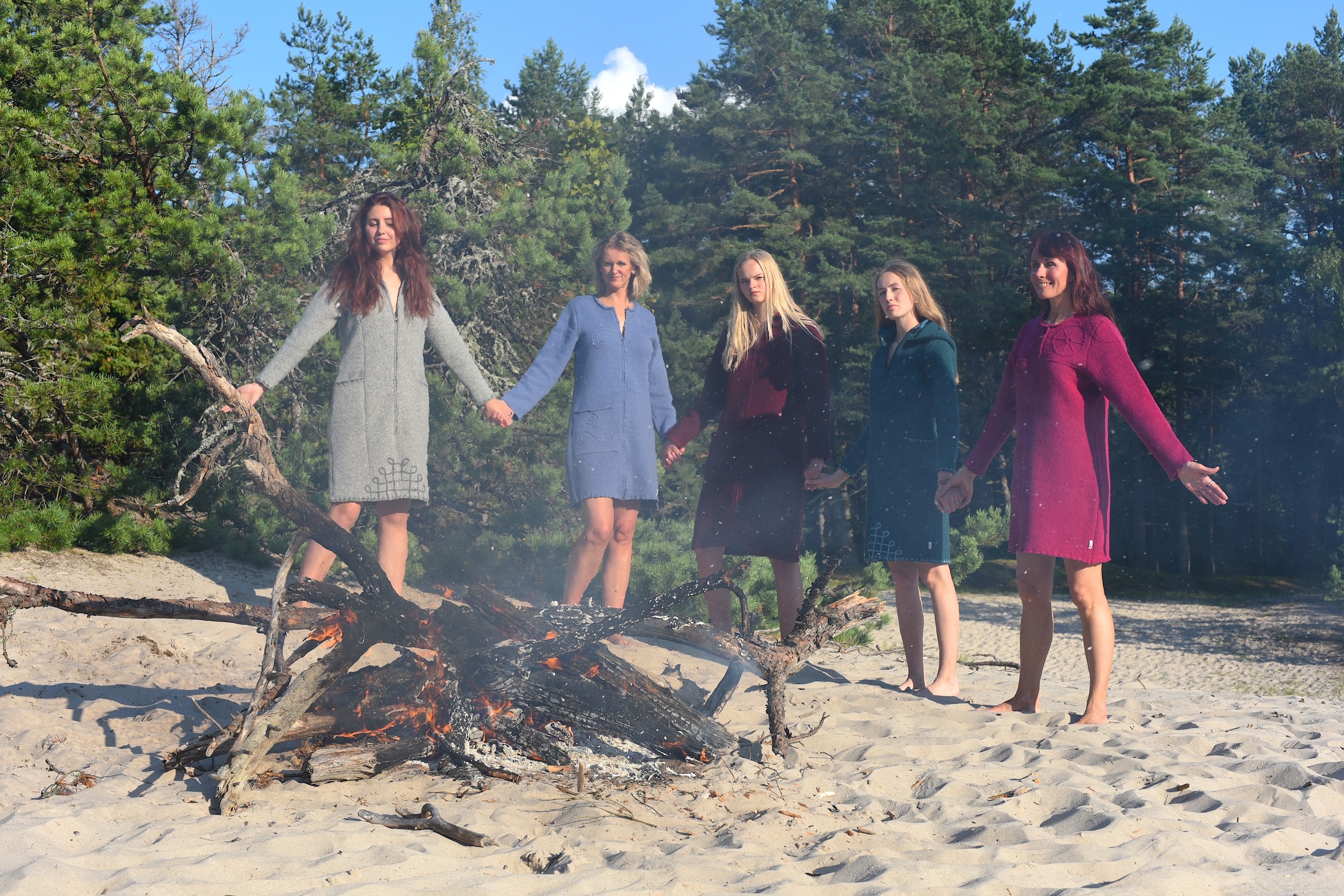 5 beautiful women in different coloured pure wool coats holding hands in front of some pine trees and a big fire