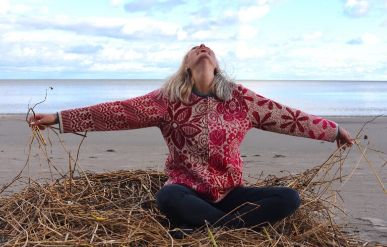 woman sitting cross legged on top of hay on a sunny beach, looking up and smiling, and wearing a graphic wool cardigan with pink flowers