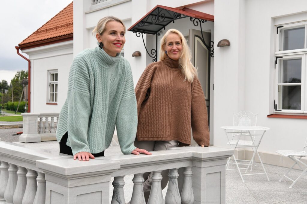 Two women in Kauni T-713 oversized braided turtleneck poncho sweaters, sage green and brown, leaning on a white manor balustrade
