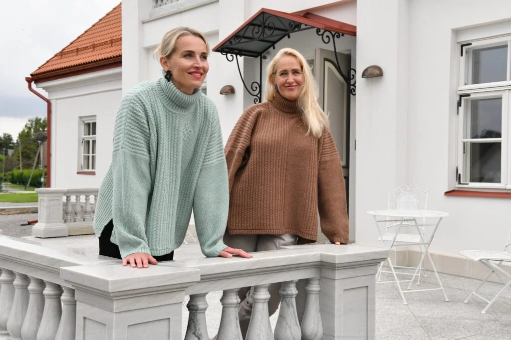 Two women in Kauni T-713 oversized braided turtleneck poncho sweaters, sage green and brown, leaning on a white manor balustrade