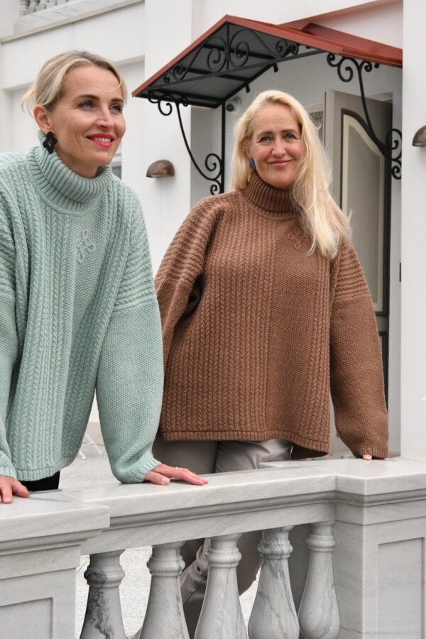 Two women in Kauni T-713 oversized braided turtleneck poncho sweaters, sage green and brown, leaning on a white manor balustrade