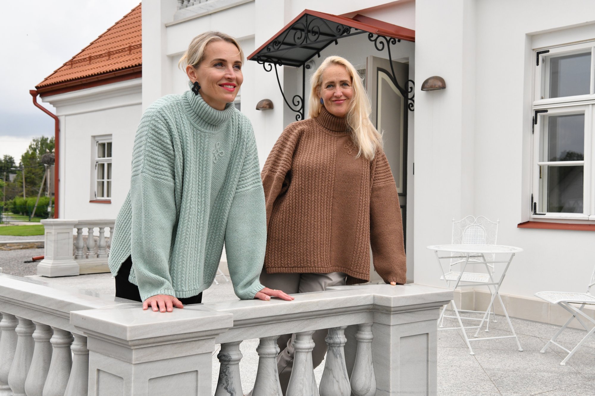 T-713 Oversized Braided Turtleneck Poncho Sweater – Sage Green Two women in Kauni T-713 oversized braided turtleneck poncho sweaters, sage green and brown, leaning on a white manor balustrade