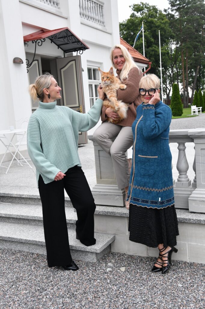 Woman in a sage green Kauni T-713 oversized braided turtleneck poncho sweater with side slits, posing on manor steps with friends and a cat